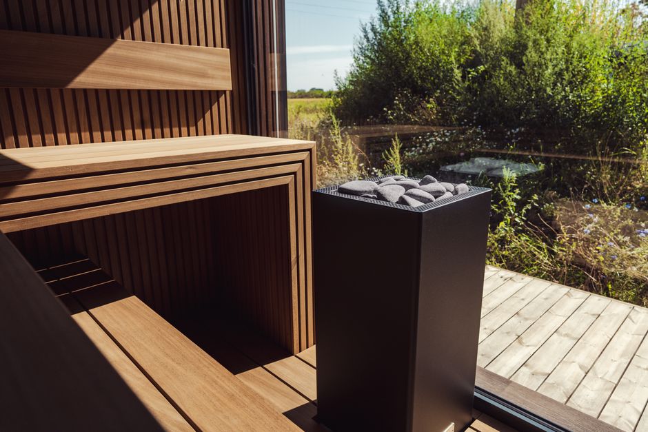 A tall dark box-shaped sauna heater topped with stones beside a bench and a glass wall