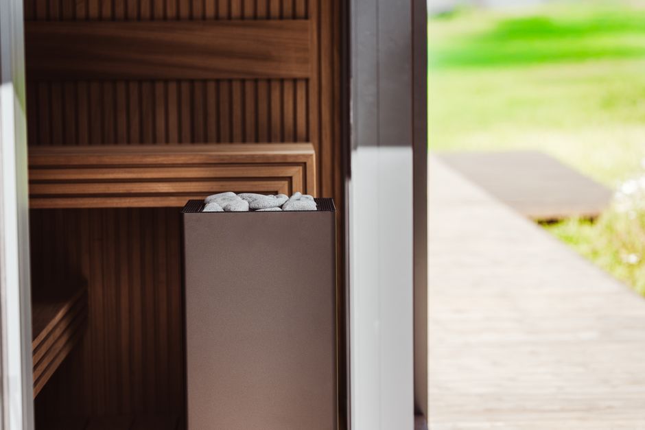 A tall grey box-shaped sauna heater topped with stones beside a wooden bench